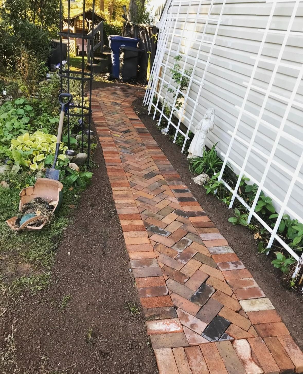 Brick garden path beside white fence, bordered by soil and plants, leading to a blue bin in the distance.
