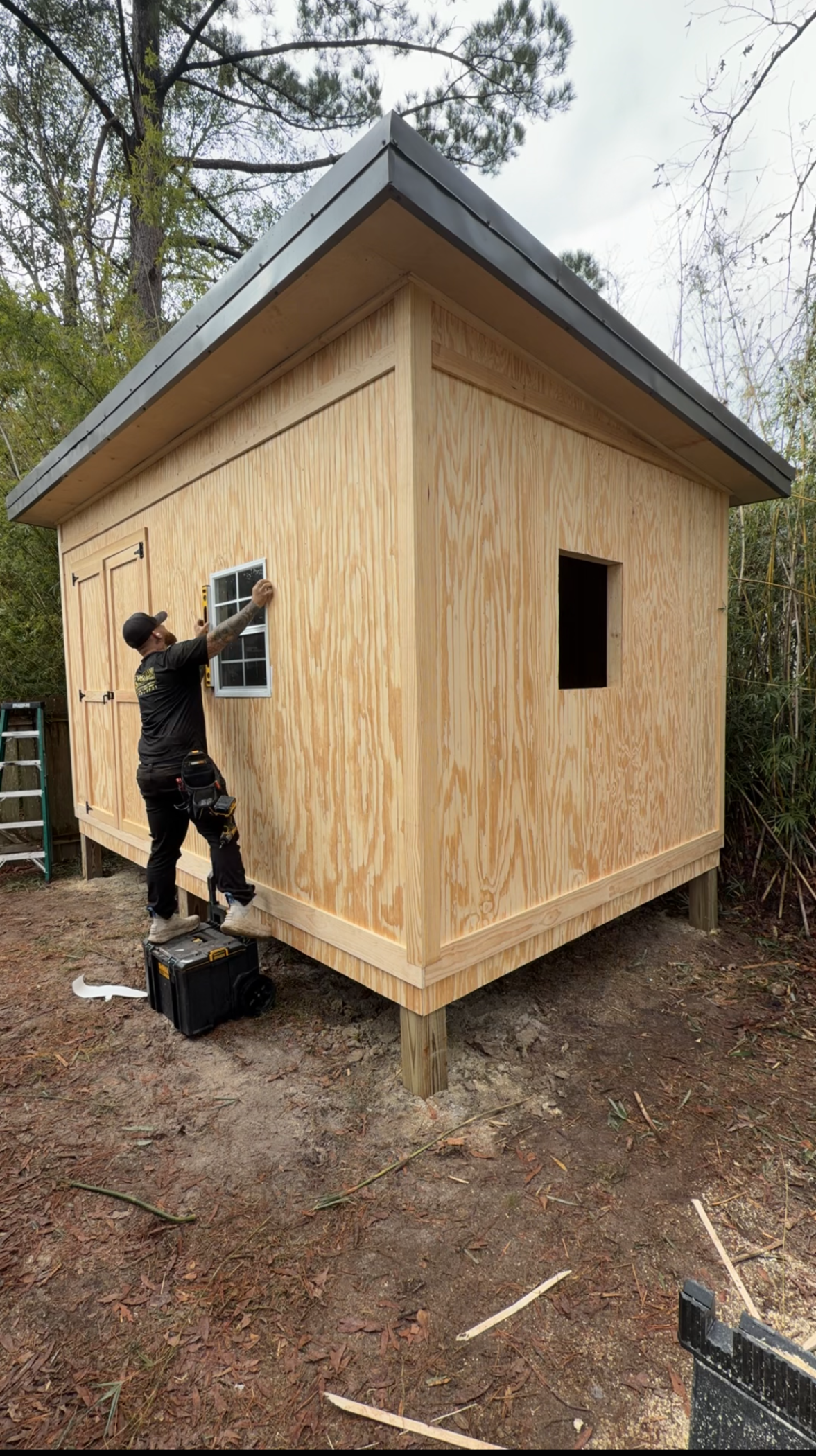 Person painting the exterior of a small wooden cabin with a dark roof in a wooded area.
