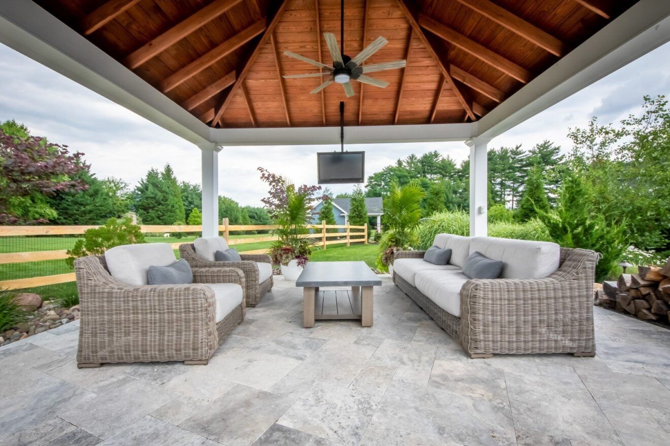 Covered patio with wicker seating, stone floor, and a wooden vaulted ceiling overlooking a green yard