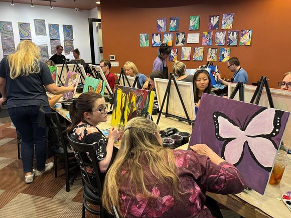 People painting at tables in an art studio, with colorful canvases and a butterfly painting in the foreground.