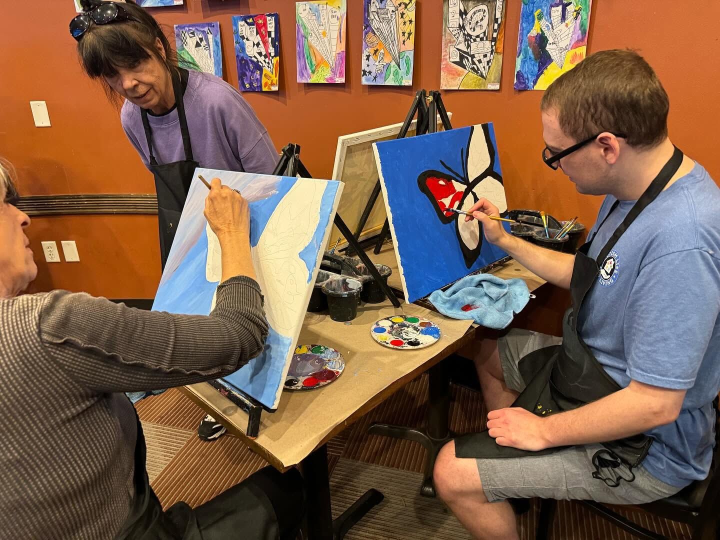 Three people paint at a table in an art studio, with colorful artwork on the wall.