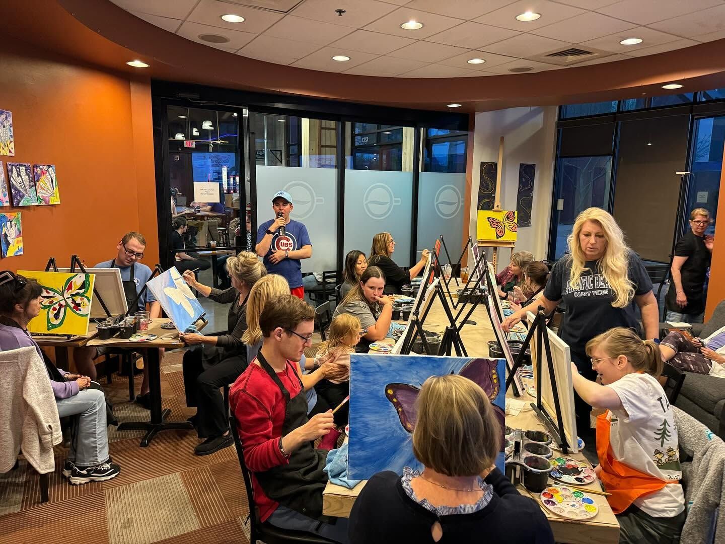 Art class in a café, with people painting at tables and a woman standing nearby.