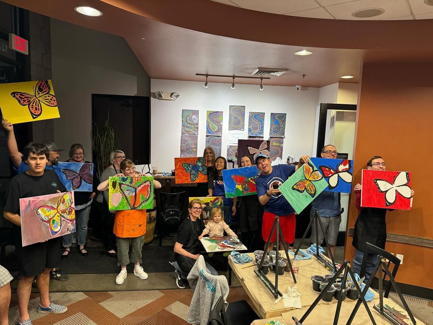 People holding colorful paintings in an art studio, posing around a table and easels