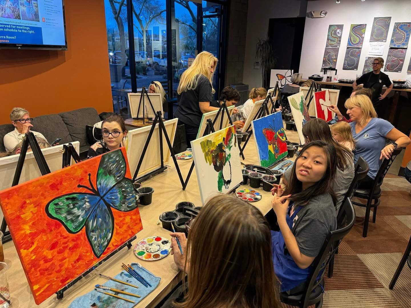People painting colorful butterfly canvases at tables in a studio class