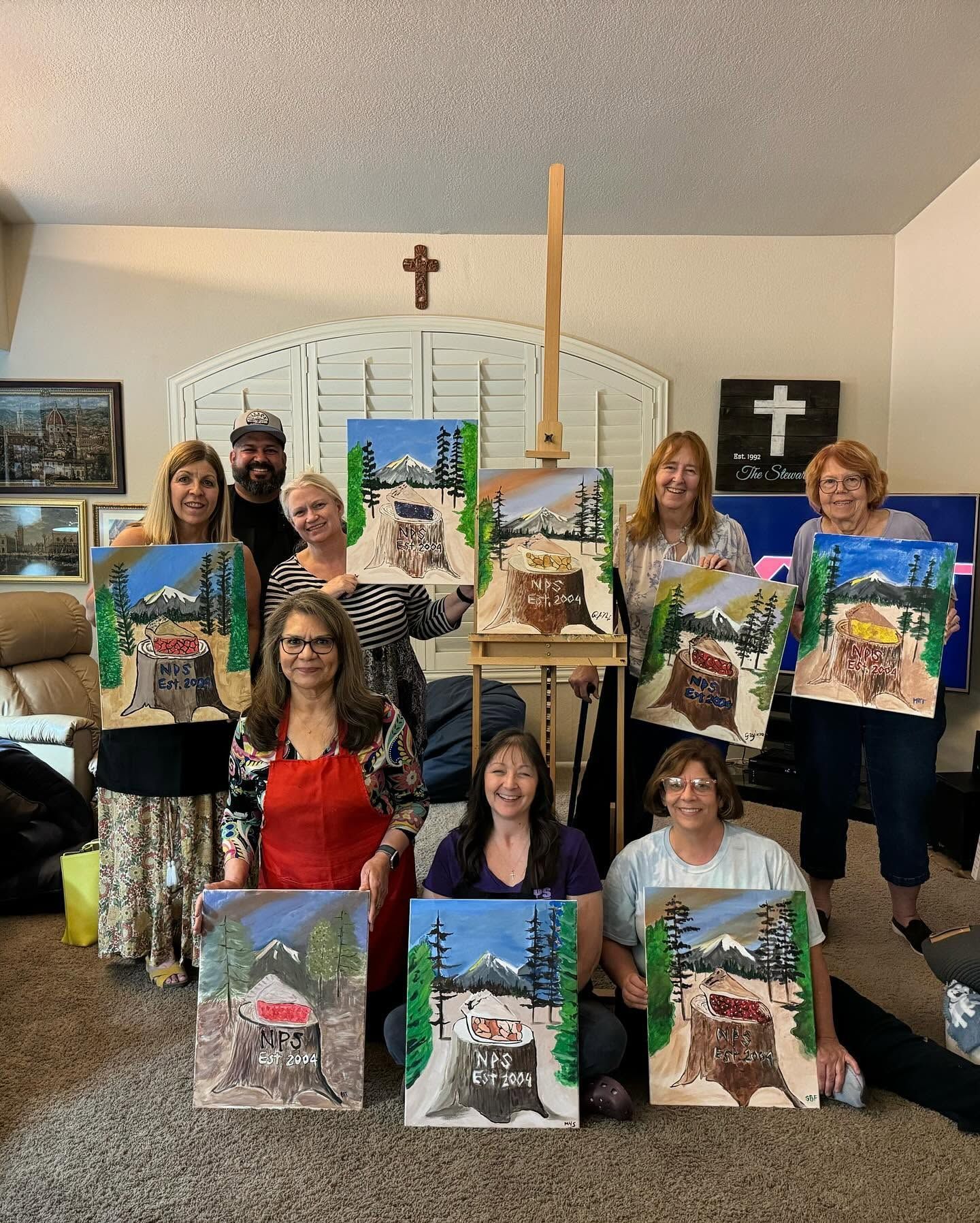 Group of people holding colorful landscape paintings in a living room, with a cross on the wall.