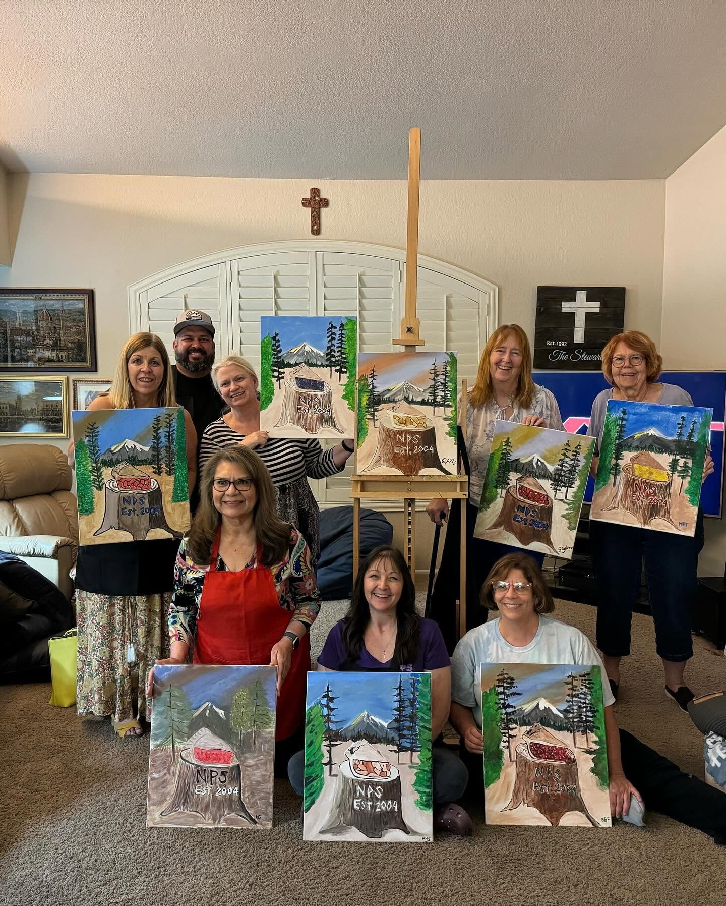 Group holds finished landscape paintings in a room with a cross on the wall.