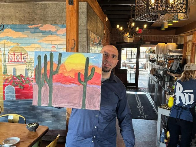 Person in a restaurant holding a colorful desert painting with cactus and sunset hues