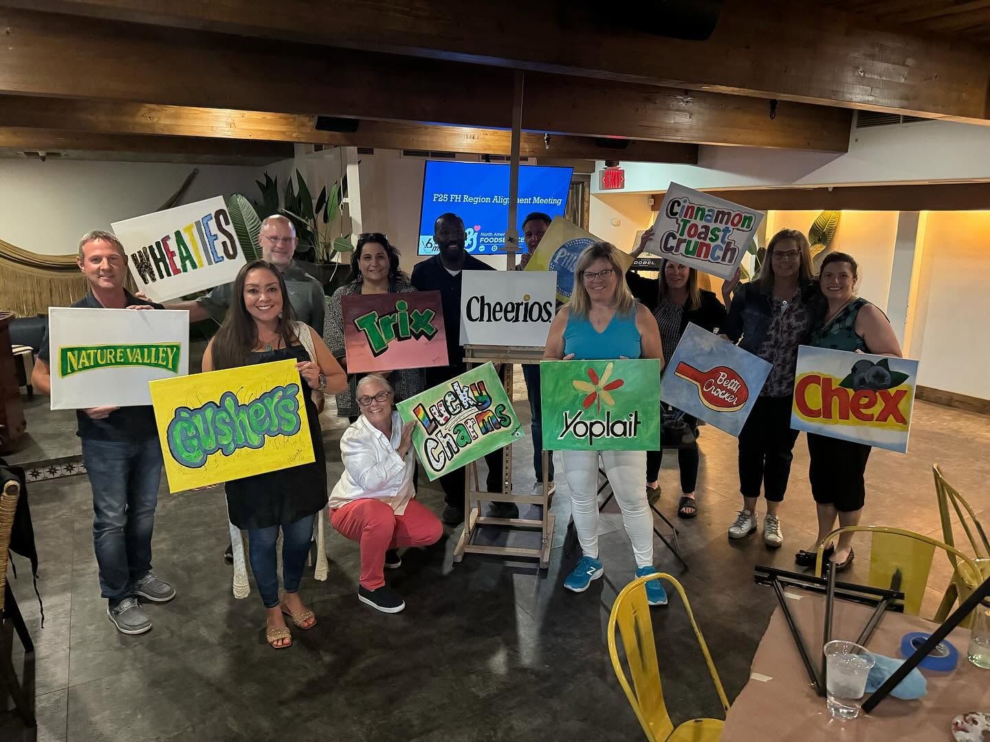 People holding colorful cereal-box signs in a casual indoor group photo.