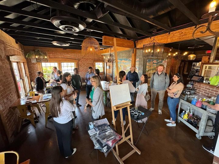 People gathered in a rustic workshop around an easel and art supplies, with brick walls and wooden beams.