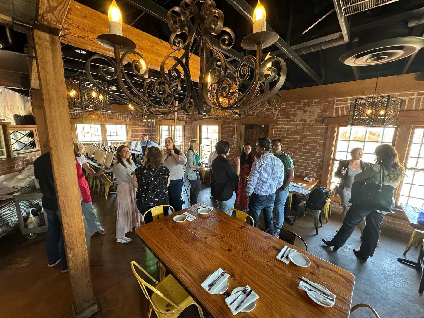 People mingling in a rustic wooden room with chandeliers and a long table set with napkins.