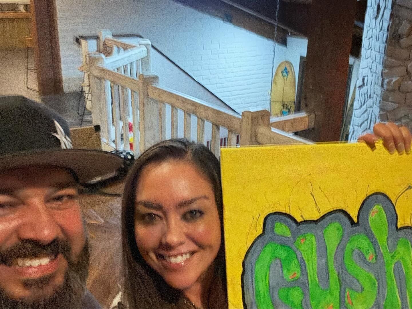 Two smiling people posing indoors beside a yellow sign with green graffiti lettering