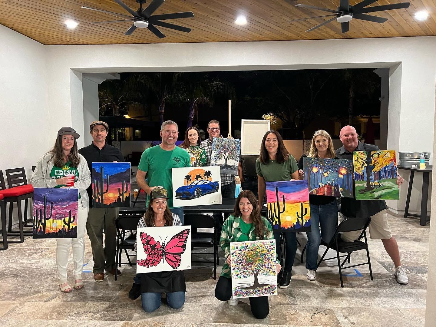 Group holding colorful sunset and butterfly paintings indoors under a pavilion with string lights