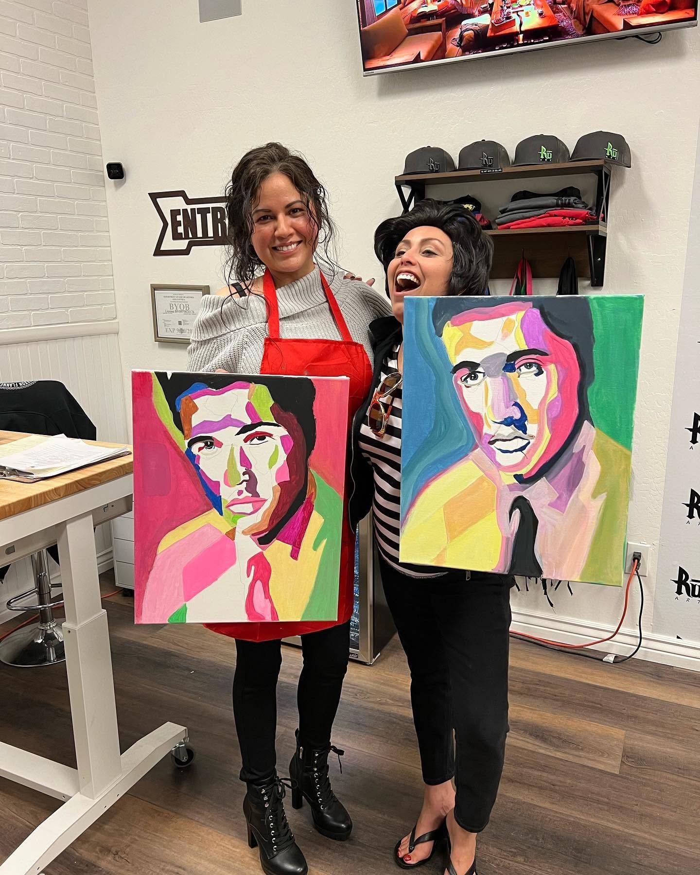 Two people hold colorful pop-art portraits in a bright studio, smiling beside an easel.