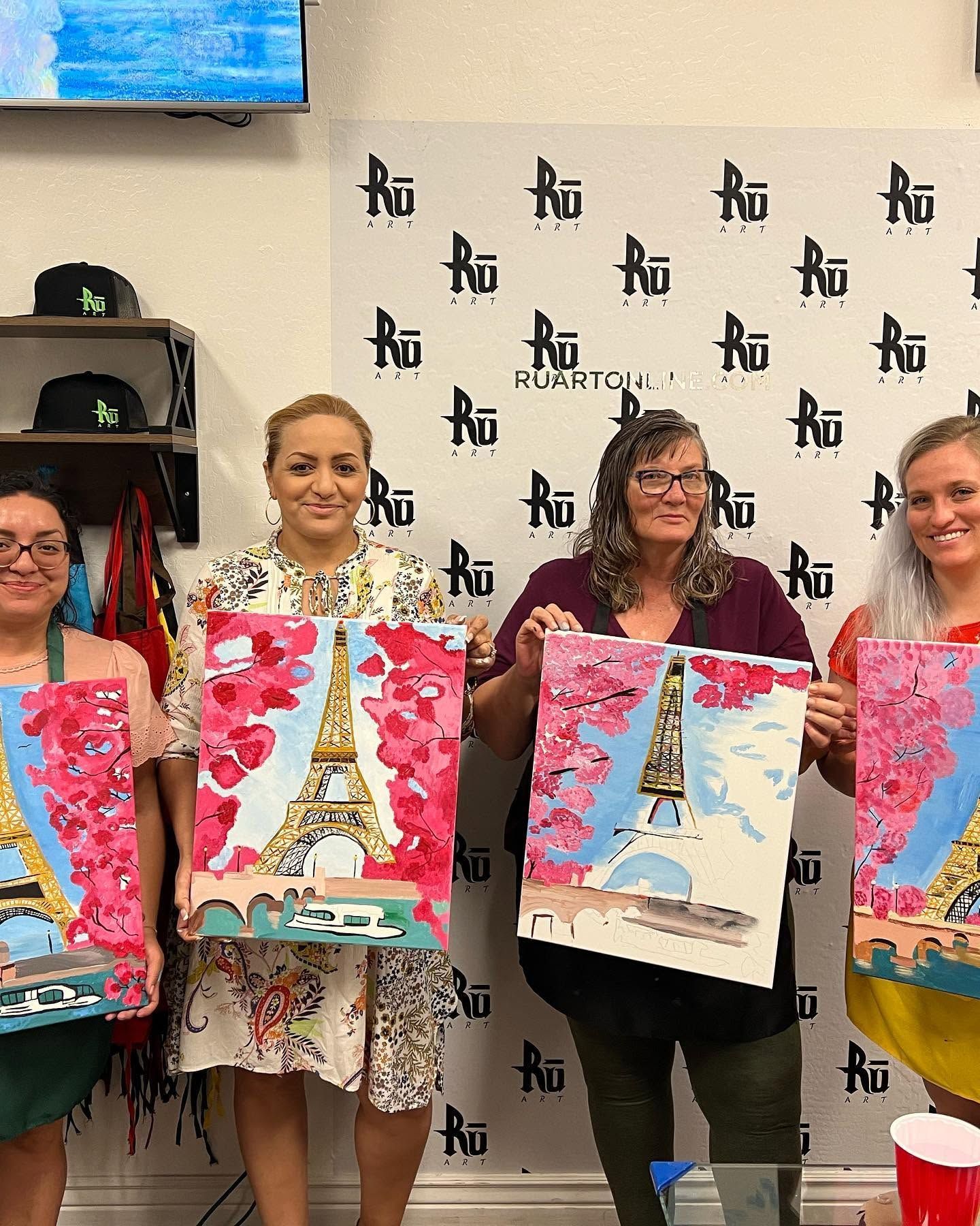 Four people holding colorful Eiffel Tower paintings in front of a photo backdrop