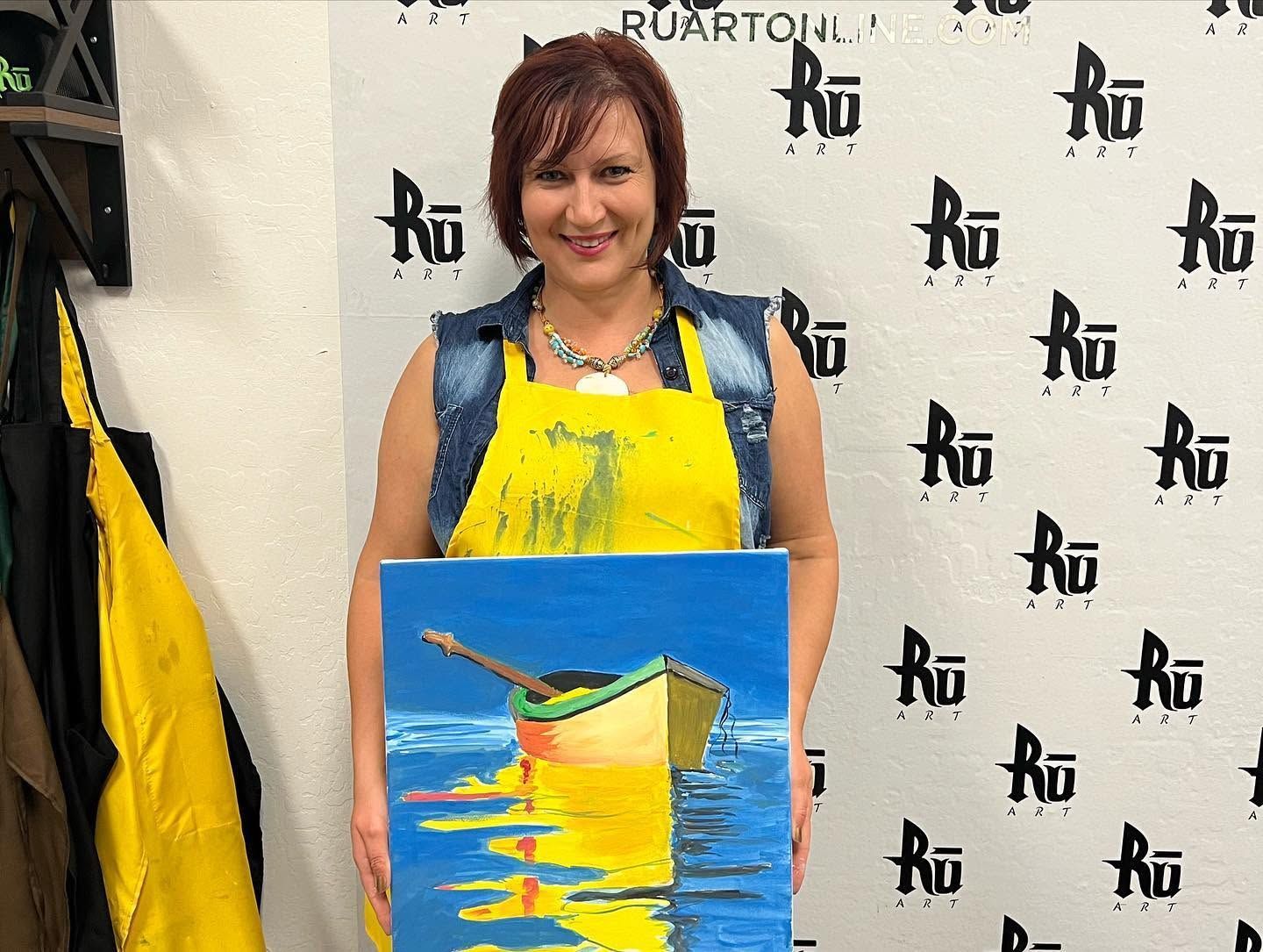 Woman in yellow apron holding a colorful painting in front of a branded backdrop