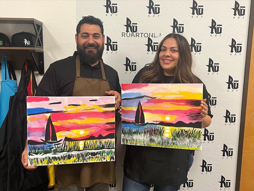 Two people holding colorful sunset paintings in front of a Royal Car Wash backdrop