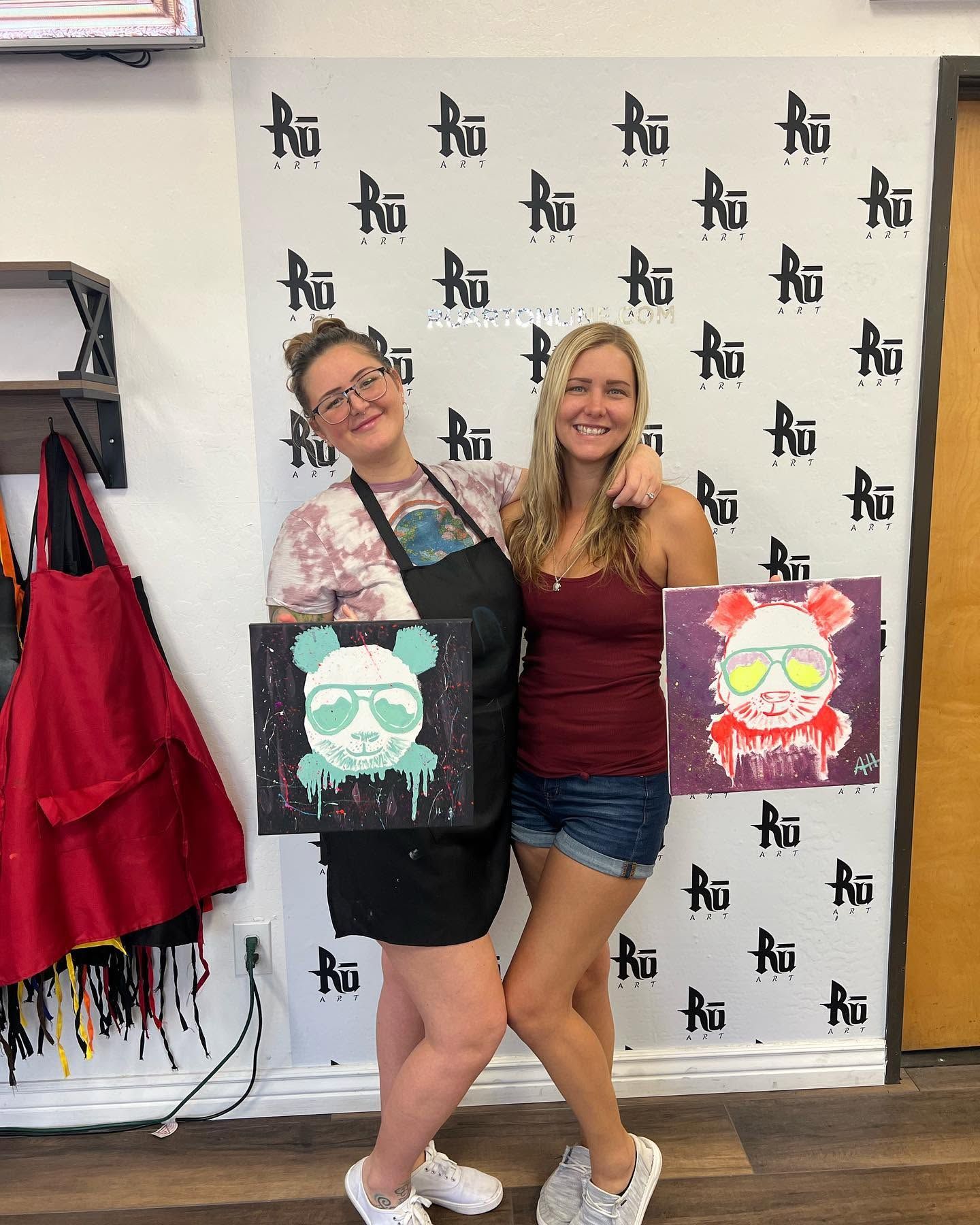 Two people posing indoors with painted tote bags in front of a branded backdrop.