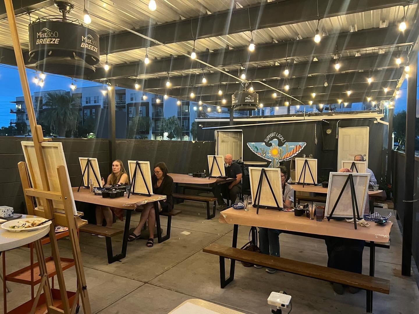Art studio with people painting at tables under string lights at dusk