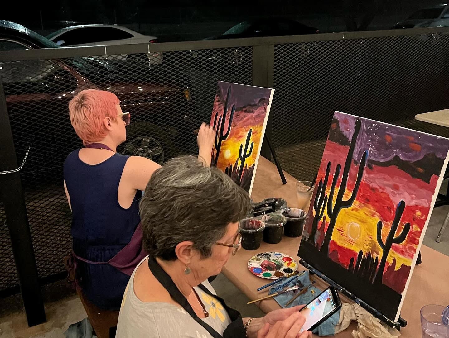 Two people paint cactus sunset scenes at easels in an outdoor art class