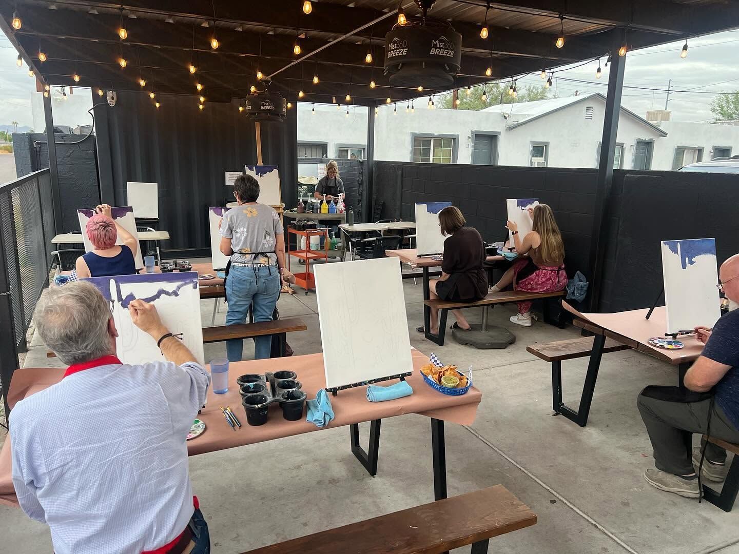 Outdoor art class under string lights, with people seated at easels and painting on a patio.