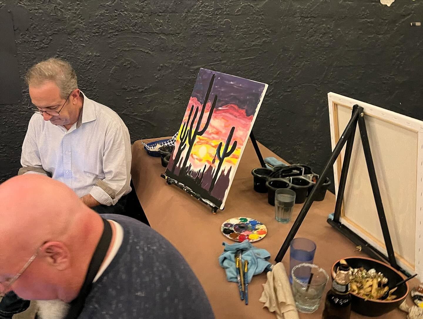 Two people painting at a table with canvases, brushes, and paint supplies in an art studio