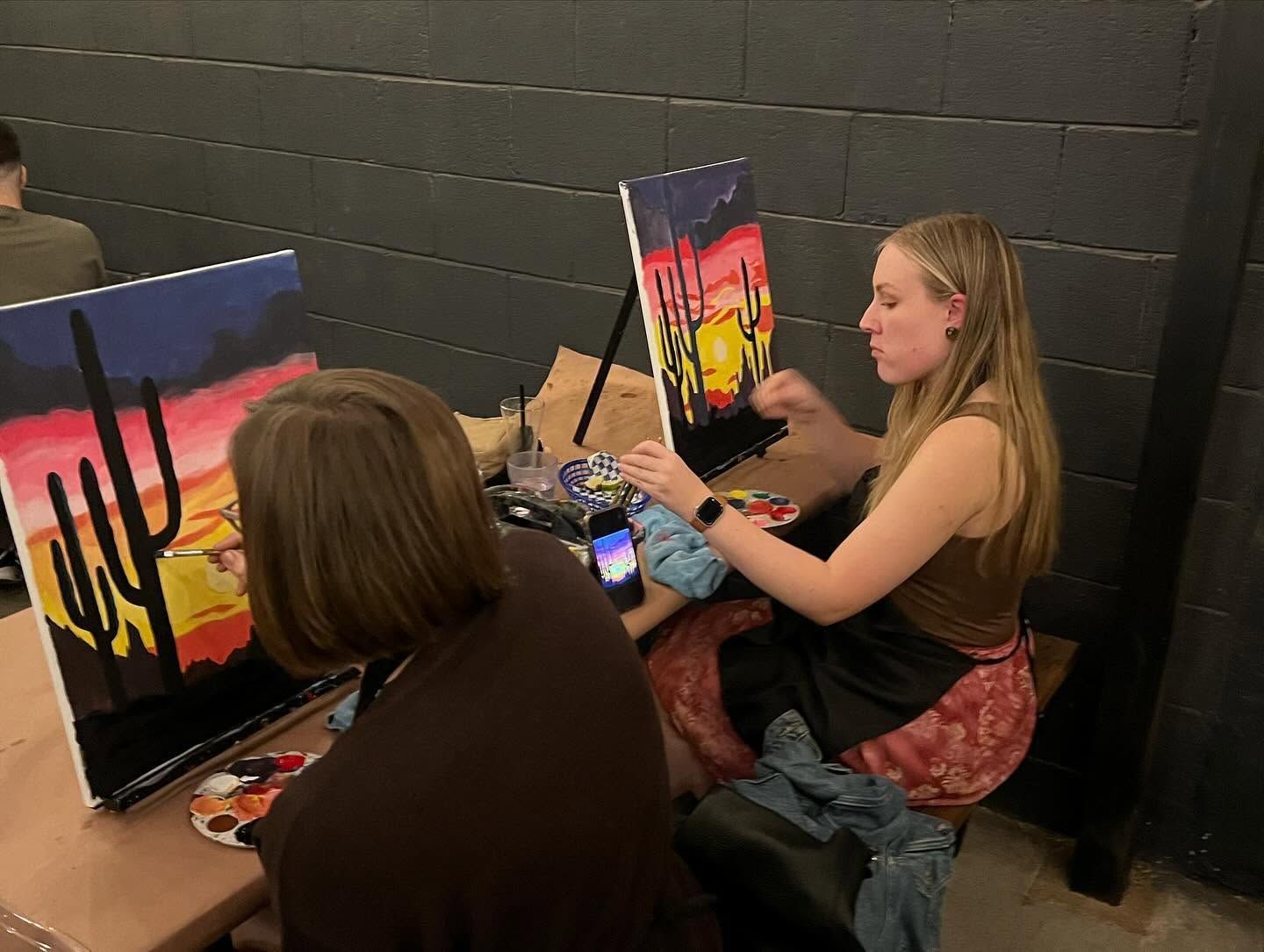 People painting colorful desert landscapes on easels in a classroom setting