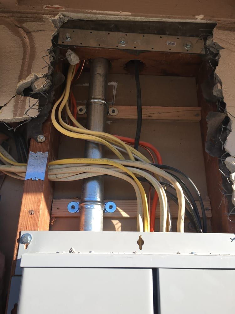 Open electrical panel with exposed wiring and a breaker box beneath a cabinet