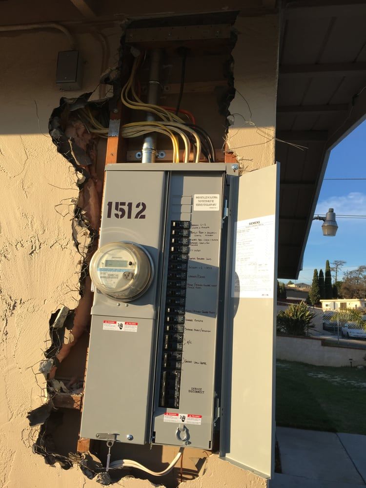 Open electrical panel on cracked exterior wall, with exposed wiring and meter box.