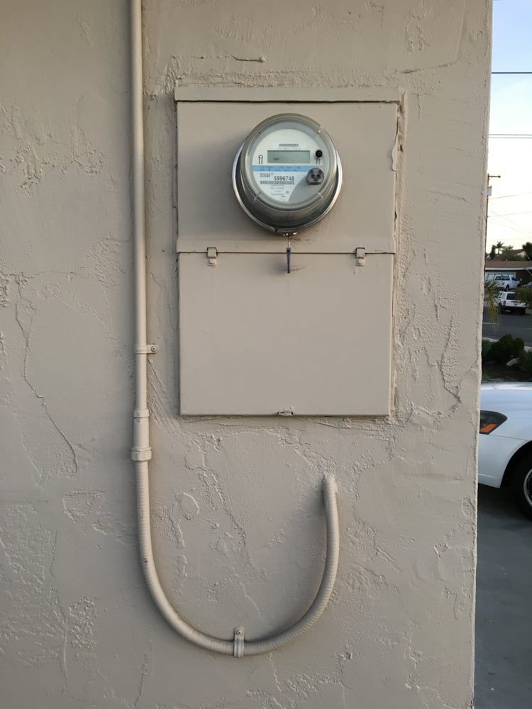 Electric meter and utility boxes on a beige exterior wall with conduit piping.