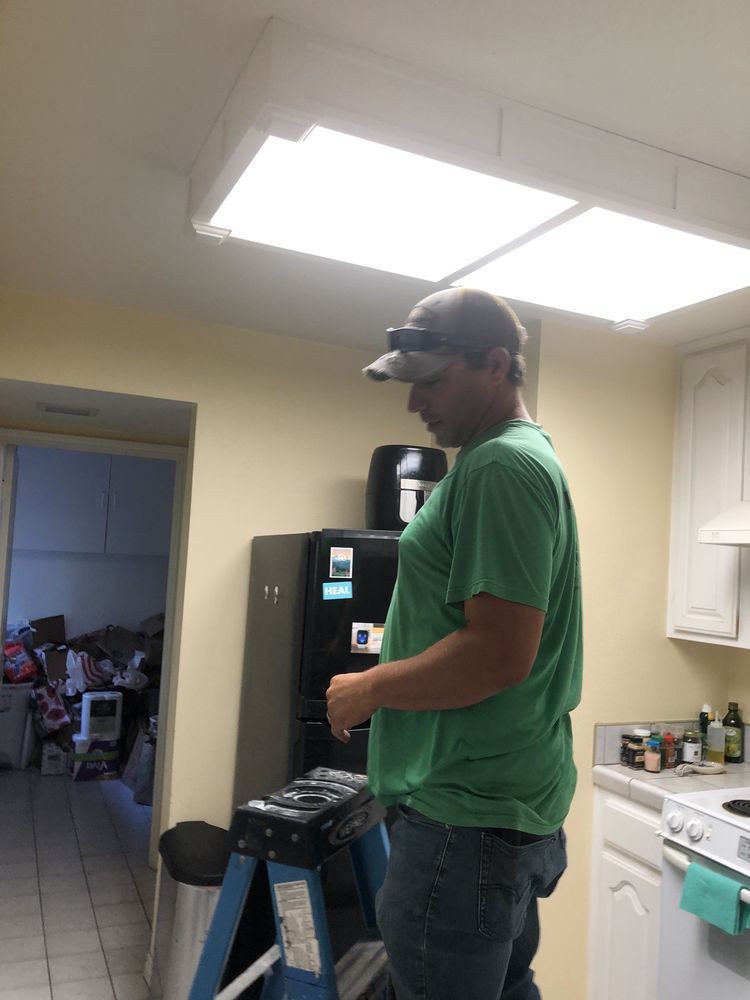 Man standing in a kitchen beside a ladder, wearing a green shirt and cap, under bright ceiling lights