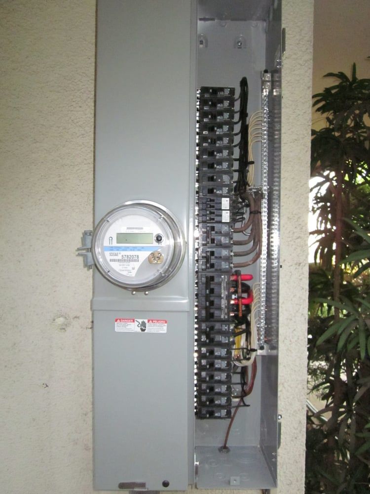 Open electrical breaker panel with meter on a wall, showing rows of circuit breakers and wiring.