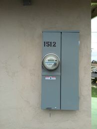 Gray electrical meter box mounted on a wall, labeled 1512, with a round meter gauge.