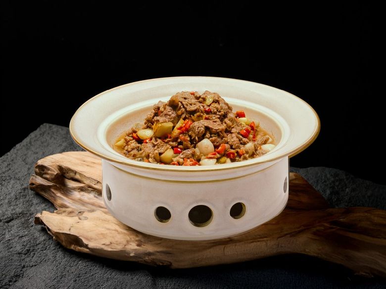 A bowl of stir-fried meat and vegetables served in a warm white ceramic pot on a textured wood base against a black backdrop.