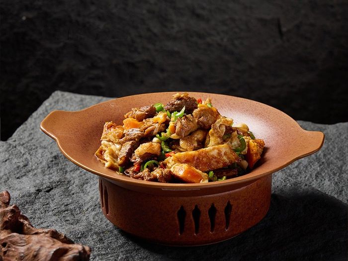 A shallow, dark orange terracotta bowl filled with a stir-fry of meat and vegetables, set on a rough, dark stone surface.