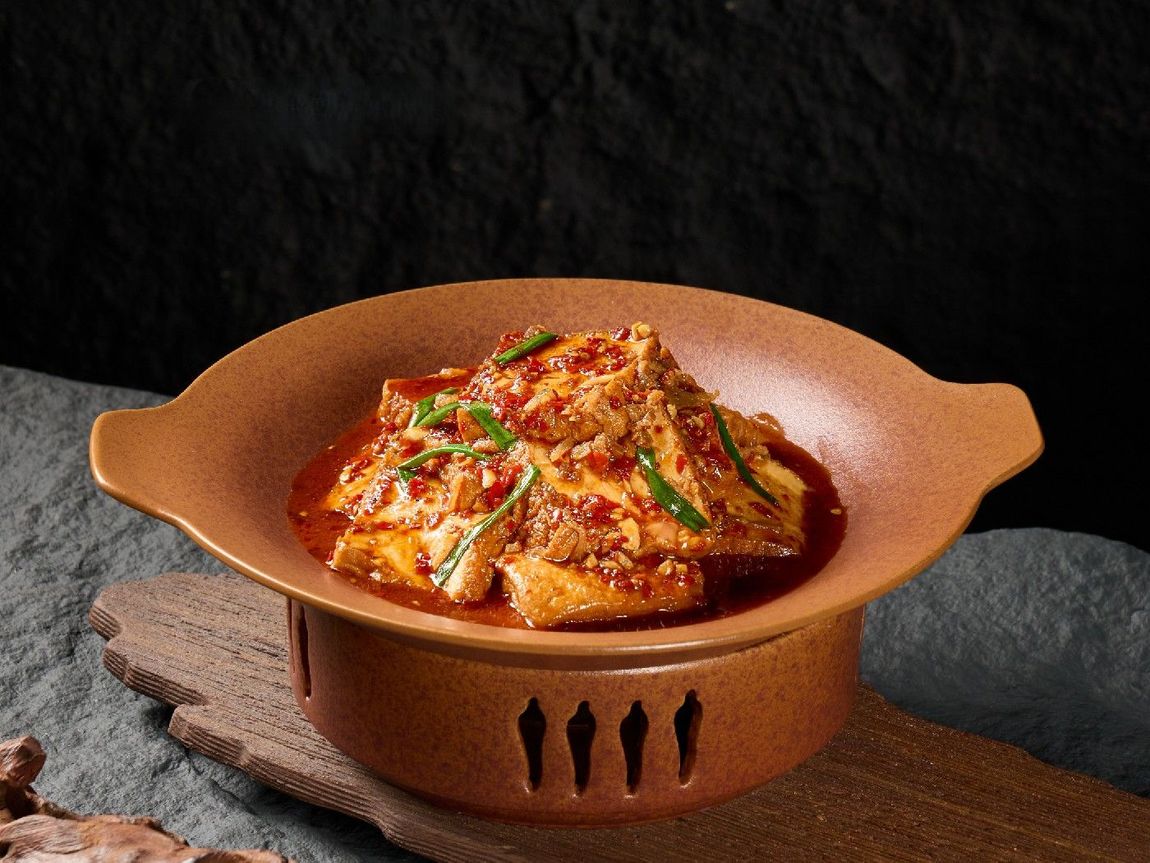 Spicy tofu dish with chopped green onions served in a warm brown clay pot on a rustic, textured dark stone surface.