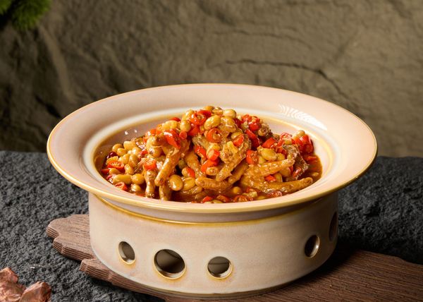 A light ceramic bowl filled with a spicy chicken feet stew featuring red peppers and yellow beans, set on a dark surface.