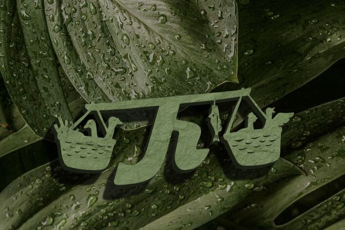 A green, cutout logo of a carrying pole with two baskets, centered on a wet, dark green leaf background.