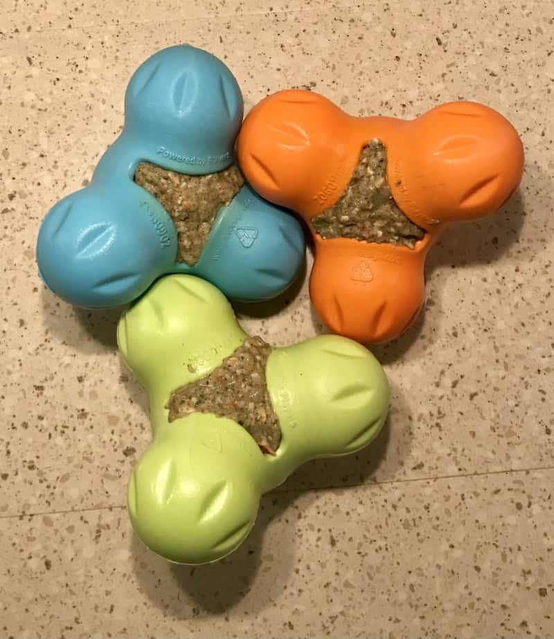 Three colorful dog toys, blue, orange, and green, filled with a treat, on a light-colored surface.