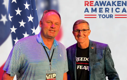 Two men posing at a Reawaken America Tour event, with an American flag backdrop and a “Freedom over fear” shirt