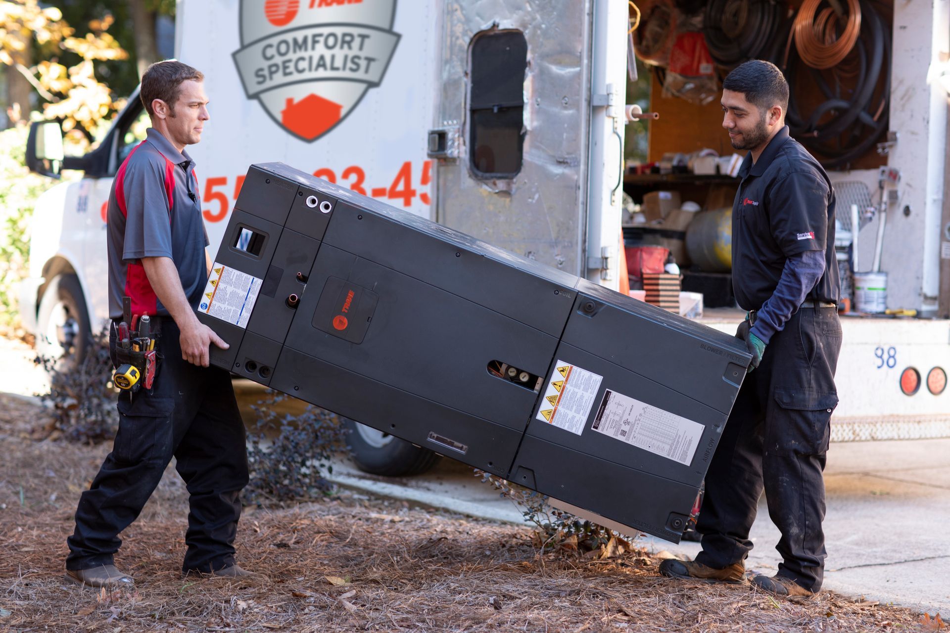 Two workers carrying a large black equipment cabinet in a wet outdoor work area