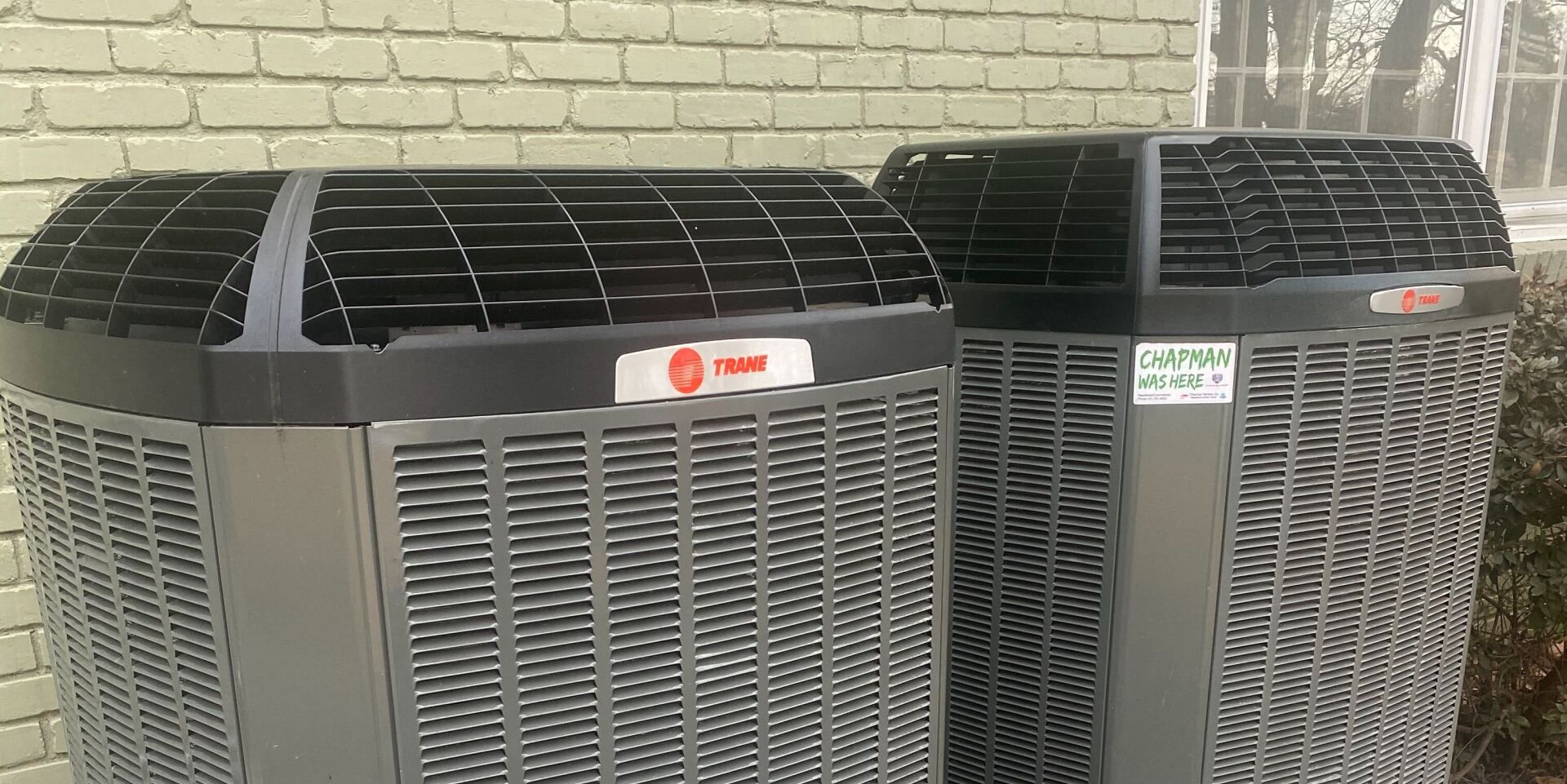 Two large outdoor air conditioning units beside a brick wall, with vents and brand labels visible.