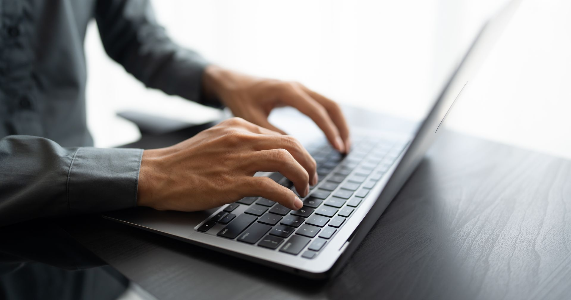Hands typing on a laptop on a dark desk, with a bright window in the background
