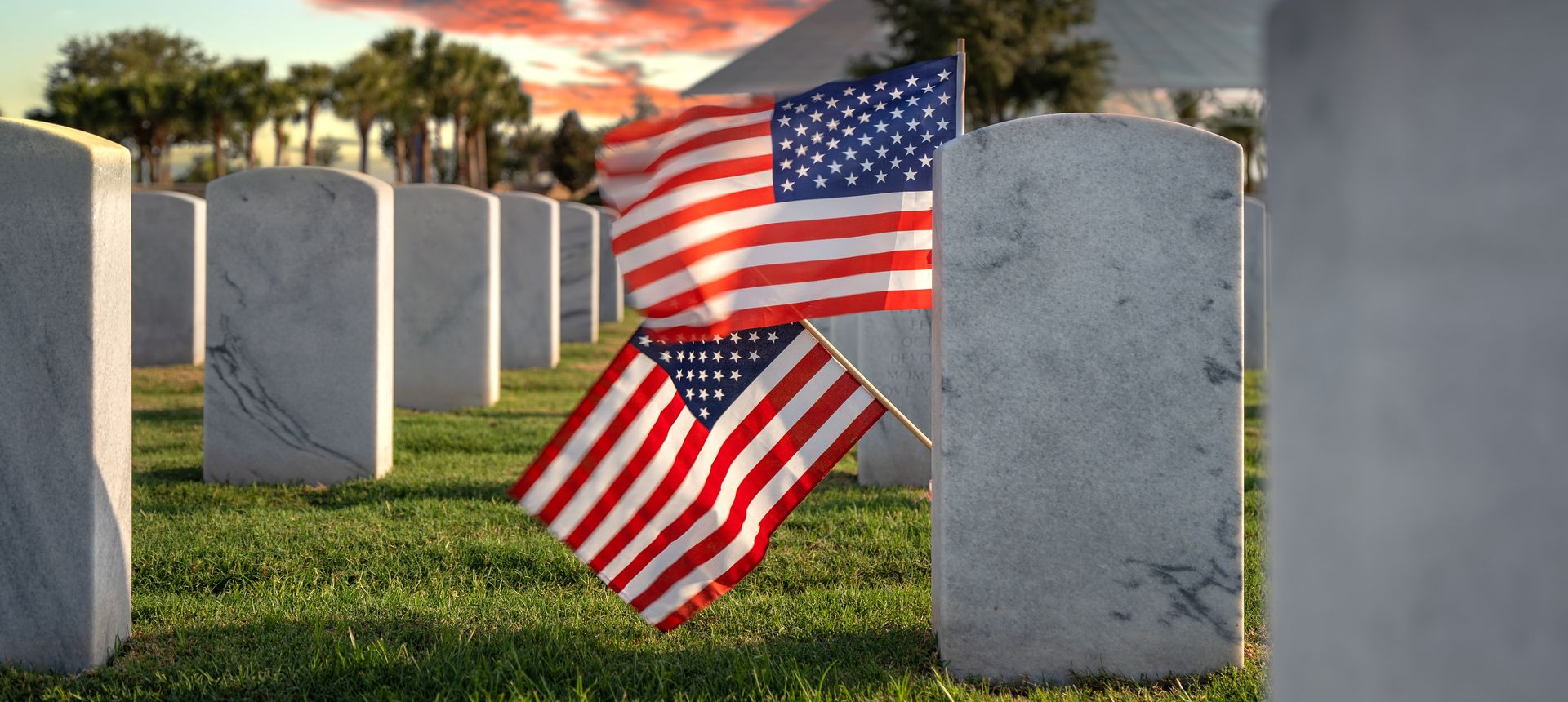 American flags placed among gravestones in a cemetery at sunset.