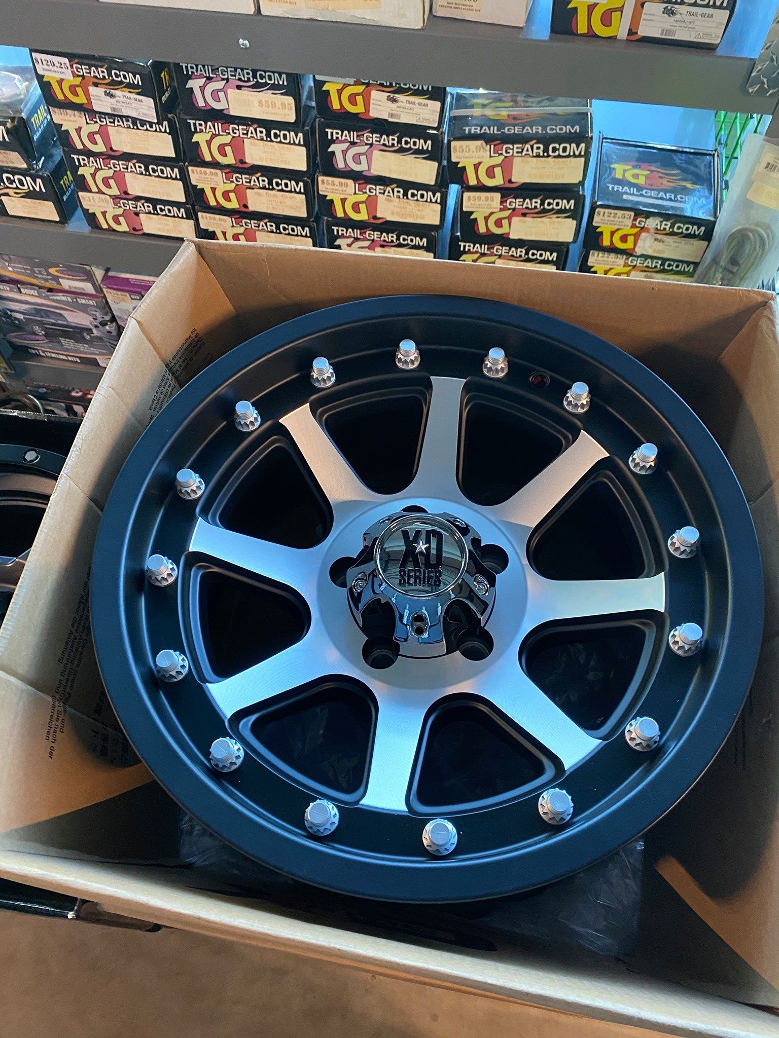 Black and silver alloy wheel with lug nuts, in a cardboard box, with shelves of boxes in the background.