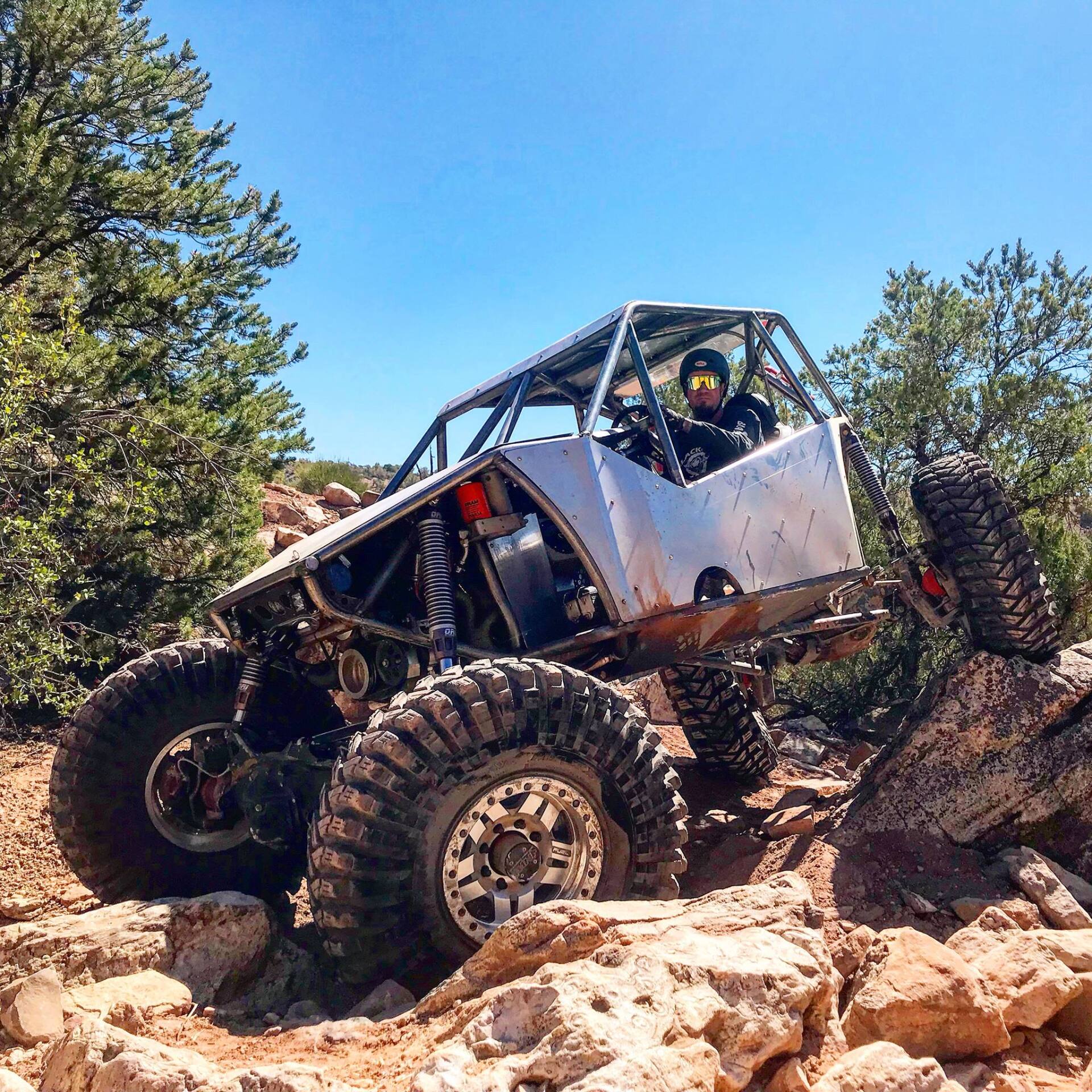 Off-road vehicle navigating rocky terrain; driver in roll cage, sunny day.