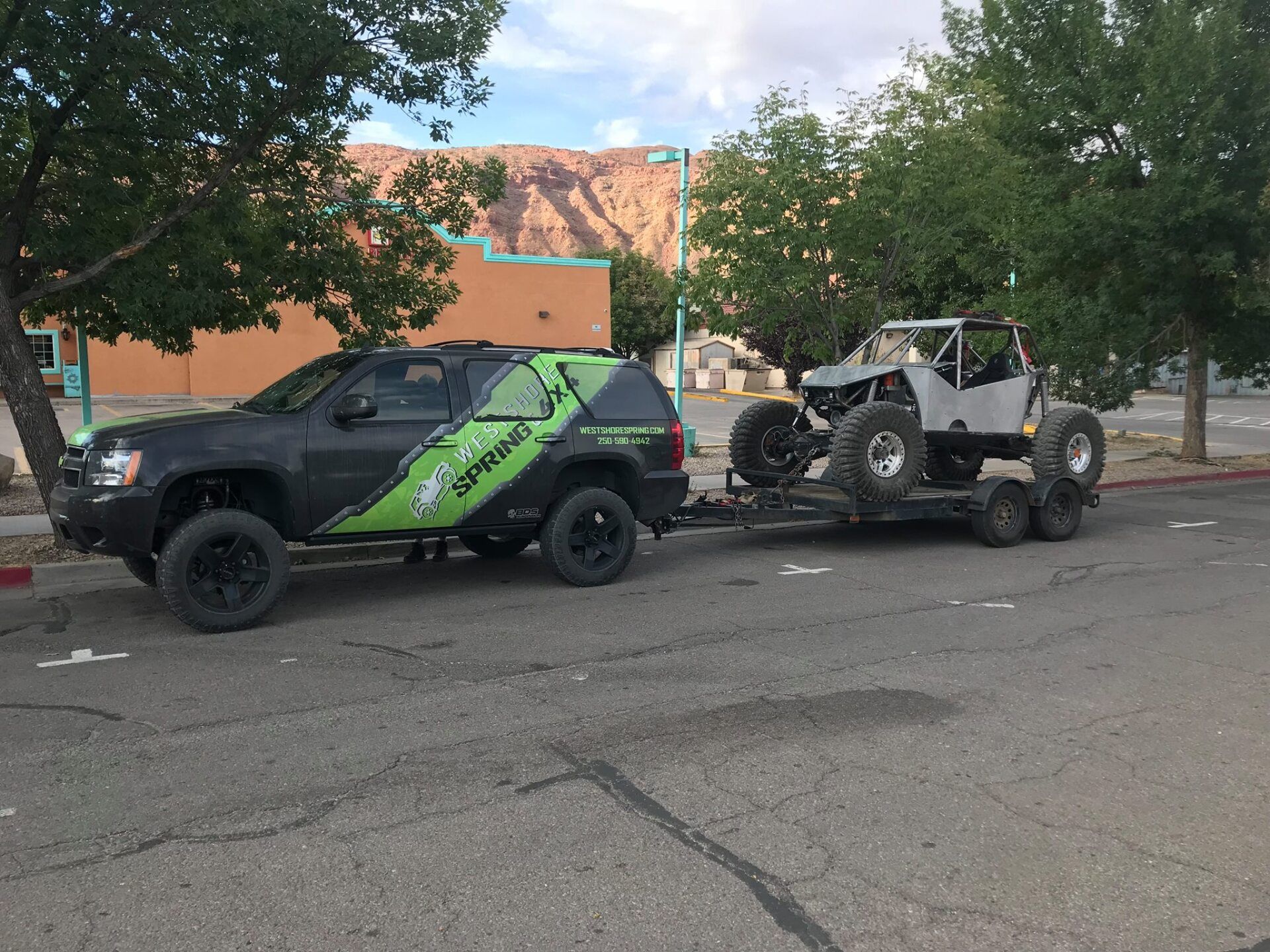 Black SUV towing a trailer with a grey off-road vehicle. Parked near trees and a building.