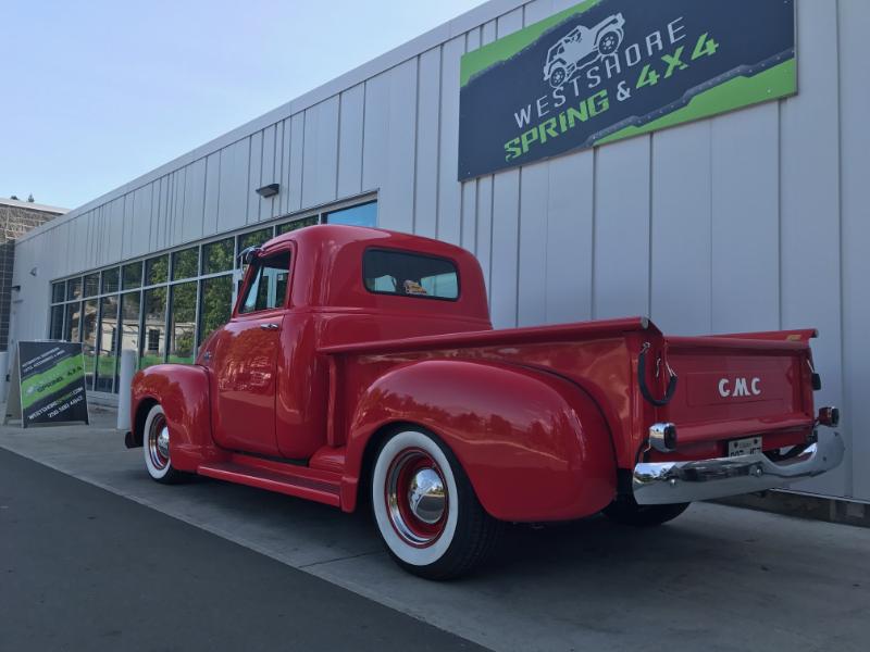 Red vintage GMC pickup truck parked in front of Westshore Spring & 4x4. White-wall tires, sunny day.