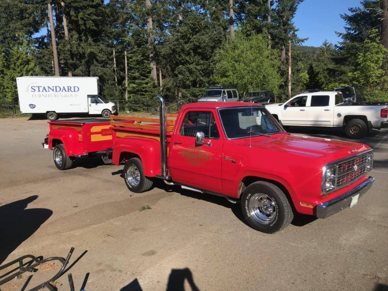 Red Dodge pickup truck towing a matching trailer. Bright sunlight, parked in front of trees, other vehicles present.