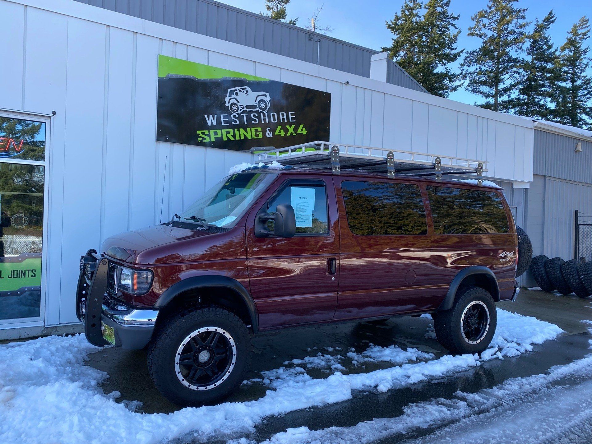 Maroon lifted Ford van with roof rack, parked in front of Westshore Spring 4x4 shop on snowy ground.