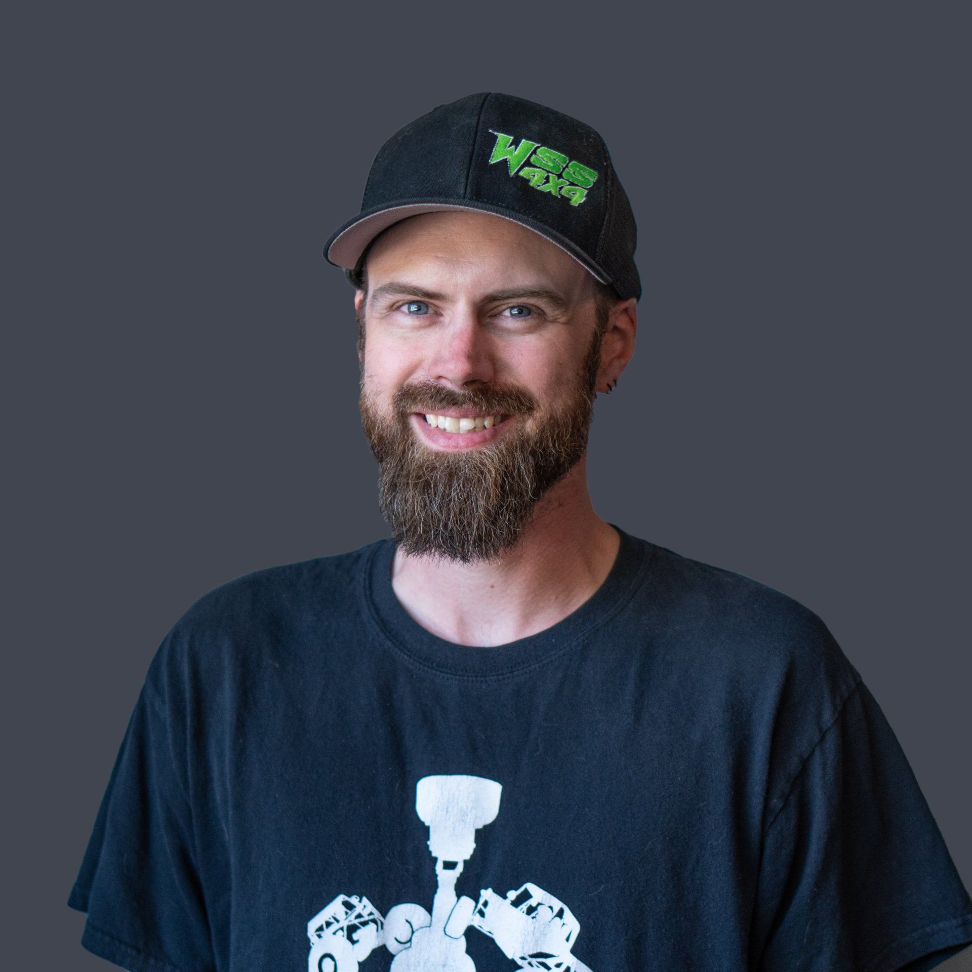 Man with beard and hat smiles. He wears a black t-shirt and hat with green logo; gray background.